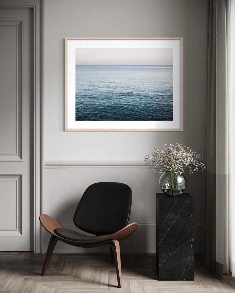 May include: A framed print of the ocean with a light wood frame. The water is a deep blue, and the sky is a soft pink and blue. A black chair with a wooden frame and a black marble pedestal with a glass vase of flowers are also in the image.