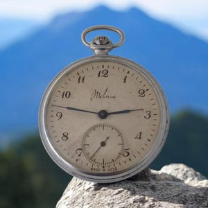 Op de afbeelding: Een vintage Molnia zakhorloge met een zilverkleurige kast en een crèmekleurige wijzerplaat. Het heeft zwarte wijzers, Arabische cijfers en een subdial. De tekst "Molnia" en "MADE IN USSR" zijn zichtbaar.
