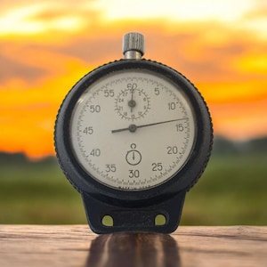 May include: A black stopwatch with a white face and a silver winding knob. The face displays numbers and markings for seconds and minutes. The background is a blurred view of a sunset.