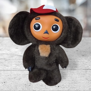 May include: A plush doll featuring a dark brown furry body with large ears, a tan chest patch, and a smooth orange face with large black eyes. The doll wears a red and white baseball cap and has a small triangular nose and a thin red mouth.