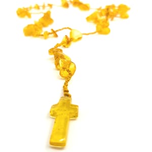 Puede incluir: Un rosario de ámbar amarillo con un colgante de cruz. El rosario está hecho de pequeñas cuentas de ámbar de forma irregular enhebradas en un cordón delgado.
