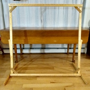 May include: A wooden loom stand with a natural finish. The stand is made of four vertical pieces of wood connected by horizontal pieces of wood. The stand is freestanding and has a rectangular shape.