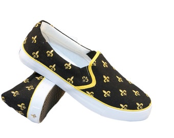 May include: Black canvas slip-on shoes with a yellow trim and a gold fleur-de-lis pattern.