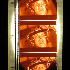 Op de afbeelding: Filmstrip met drie frames van een persoon met een metalen hoofddeksel, met ogen wijd open van angst. De filmstrip heeft het nummer "125" aan de zijkant gedrukt. De afbeelding is verlicht met een oranje tint.