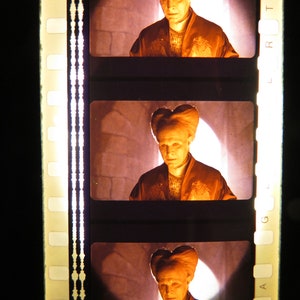 Puede incluir: Una tira de película con tres fotogramas que muestran a una persona con un peinado en forma de corazón que lleva una túnica larga y oscura. La persona está en una habitación con poca luz con una fuente de luz detrás de ella.