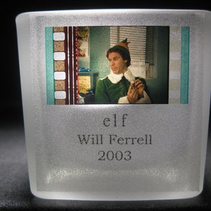 May include: A frosted glass paperweight with a film strip image of Will Ferrell as Buddy the Elf from the movie "Elf". The text "elf" and "Will Ferrell" are printed below the image, and the year "2003" is printed below that.