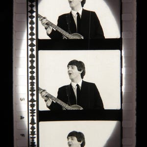 Op de afbeelding: Zwart-wit filmstrip met drie frames van een man in een pak die een basgitaar bespeelt. De man zingt en een schijnwerper schijnt op hem in elk frame. De filmstrip heeft perforaties langs de linkerrand.