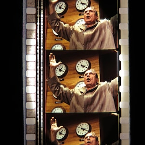 May include: A film strip featuring three frames of a man with his arms raised, set against a backdrop of clocks. The clocks display different times, and the man appears to be in a state of surprise or alarm. The film strip has a black background.
