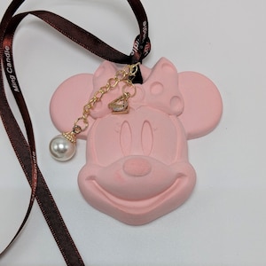 May include: Pink Minnie Mouse car air freshener with a brown ribbon and a gold chain with a pearl and a small diamond charm. The air freshener has a polka dot bow on top of its head.