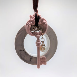 Puede incluir: Un adorno en forma de llave rosa y gris con un dije de perla que cuelga de una cadena dorada. El adorno está sujeto a una cinta marrón.