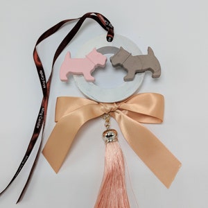May include: A white ceramic ring with two small dog figurines, one pink and one gray, attached. The ring is tied with a peach-colored ribbon and has a peach-colored tassel hanging from it. The ribbon has a brown tag with the text "Ming Candle" on it.