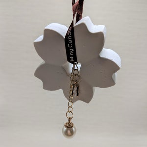 May include: White ceramic flower-shaped air freshener with a brown ribbon that says "Ming Candle" and a gold chain with a pearl hanging from it.