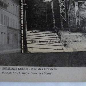 May include: Black and white photographic postcard of a street scene in Soissons, France. The text on the postcard reads "Clich&eacute; Section photographique de l'Arm&eacute;e 716 - SOISSONS (Aisne) - Rue des Graviers SOISSONS (Aisne) - Graviers Street"