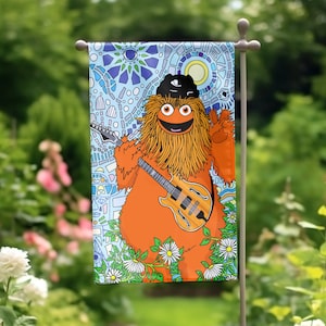 May include: A colorful garden flag featuring a cartoon orange monster with long brown hair playing a guitar. The monster is wearing a black hat and is surrounded by flowers. The background is a blue and white mosaic pattern.