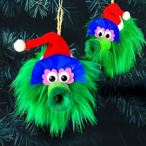 May include: Two green fuzzy Christmas ornaments with red and blue hats. Each ornament has googly eyes and a pink nose.