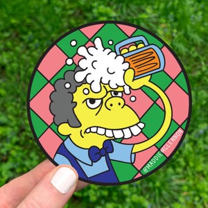 May include: A round sticker featuring a cartoon character with yellow skin, gray hair, and a blue shirt, pouring beer over his head. The background is a pink and green checkered pattern. The sticker has the text "@MADDIE MCLENNON".
