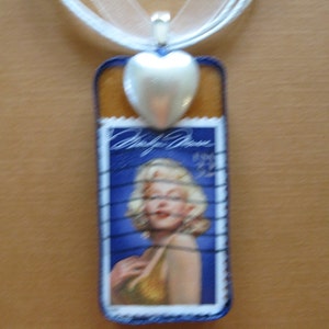 Puede incluir: Un collar con un colgante de corazón plateado y un sello postal azul y blanco con un retrato de Marilyn Monroe. El sello tiene el texto "Marilyn Monroe" y "USA".
