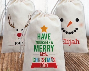 May include: Three off-white drawstring gift bags made of fabric. One bag features a reindeer face and the name JOHN. The center bag displays a yellow star and the text HAVE YOURSELF A MERRY little CHRISTMAS JOEY. The third bag shows a snowman face and the name Elijah.