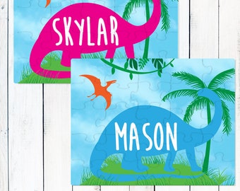 May include: Two rectangular jigsaw puzzles featuring a dinosaur theme. One puzzle displays a pink long-necked dinosaur with the name Skylar, and the other shows a blue long-necked dinosaur with the name Mason. Both include orange pterodactyls.