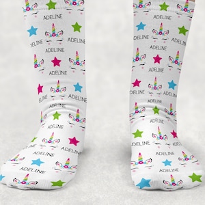 May include: A pair of white crew socks featuring a repeating pattern of unicorn faces with colorful horns, pink cheeks, and closed eyes. The design includes scattered blue, green, and pink stars, along with the name ADELINE printed in black text.