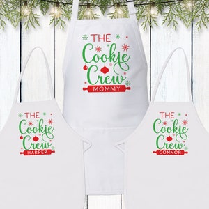 May include: Three white aprons with red and green text that reads "The Cookie Crew" with a name below each. The top apron says "Mommy", the middle apron says "Harper", and the bottom apron says "Connor".