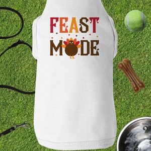 Op de afbeelding: Witte hondenvest met de tekst "FEAST MODE" in rode, oranje en bruine letters. Een cartoon-kalkoen bevindt zich in het midden van de tekst.