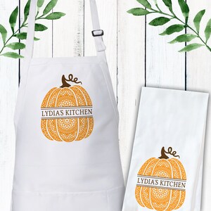 May include: A white apron and a matching white kitchen towel, both featuring an orange pumpkin graphic with a brown stem and the text LYDIA'S KITCHEN printed in brown across the centre of the pumpkin, which has a white mandala pattern.