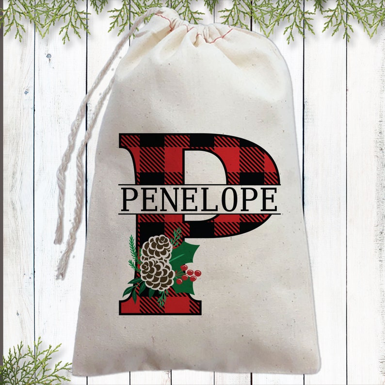 Pu&ograve; includere: Un sacchetto regalo in cotone bianco con una lettera "P" a quadri rossi e neri e il nome "PENELOPE" stampato su di esso. La lettera &egrave; decorata con una pigna e un ramo di agrifoglio.