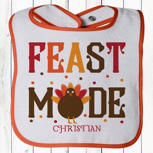 K&ouml;nnte beinhalten: Ein wei&szlig;er Baby-L&auml;tzchen mit orangefarbenem Rand. Es zeigt einen braunen Truthahn mit orangefarbenen und roten Federn und dem Text "FEAST MODE" in Rot, Orange und Braun. Der Name "CHRISTIAN" ist in Wei&szlig; unter dem Truthahn gedruckt.