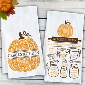 May include: Two white kitchen towels featuring brown and orange graphics. One towel displays a large pumpkin with a mandala pattern and the text Grace's Kitchen. The other shows a pumpkin, a rolling pin, and kitchen measurement conversions.