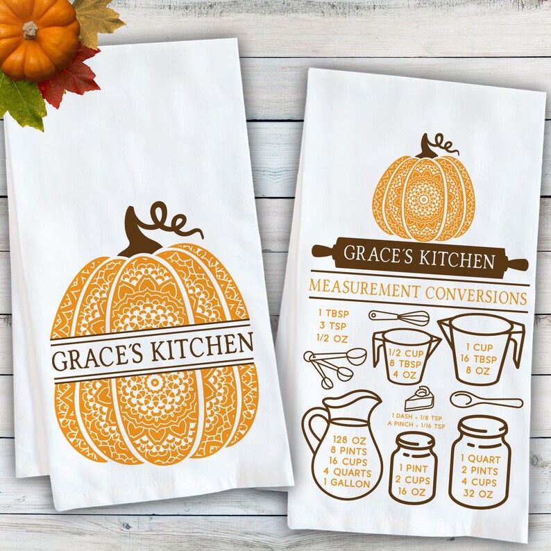 May include: Two white kitchen towels featuring brown and orange graphics. One towel displays a large pumpkin with a mandala pattern and the text Grace's Kitchen. The other shows a pumpkin, a rolling pin, and kitchen measurement conversions.