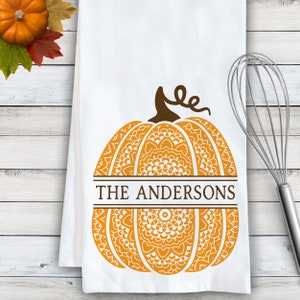 May include: A white kitchen towel featuring a large orange pumpkin graphic with intricate white mandala-style patterns. The pumpkin is bisected by a horizontal brown bar containing the text THE ANDERSONS in brown capital letters.