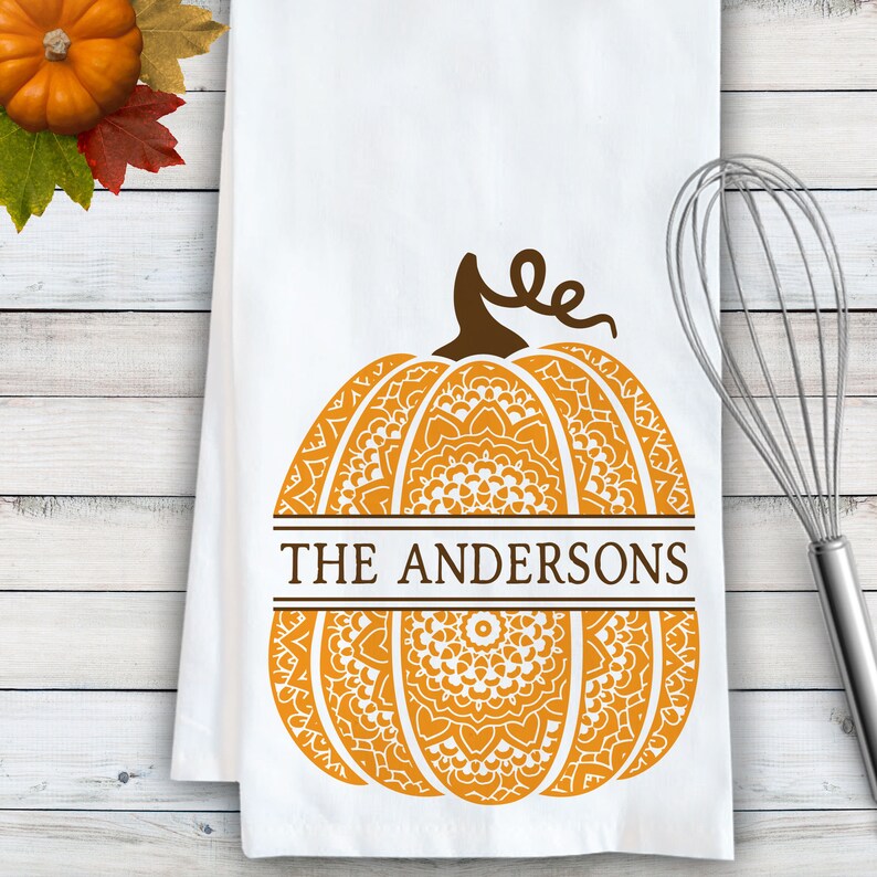 May include: A white kitchen towel featuring a large orange pumpkin graphic with intricate white mandala-style patterns. The pumpkin is bisected by a horizontal brown bar containing the text THE ANDERSONS in brown capital letters.