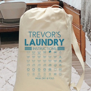 May include: A white canvas laundry bag with blue text that reads "Trevor's Laundry Instructions" and includes a chart of laundry care symbols.