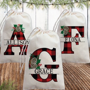 Pu&ograve; includere: Tre sacchetti regalo in tessuto bianco con lettere in plaid rosso e nero e un design a pigna e agrifoglio. I sacchetti sono personalizzati con i nomi Allison, Fiona e Grace.