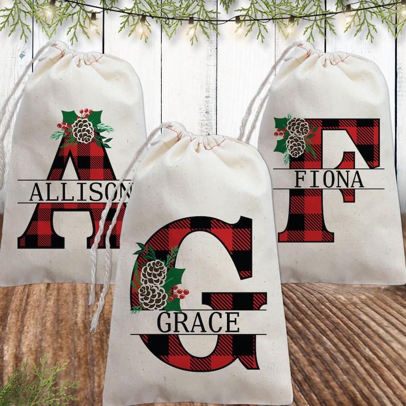 Pu&ograve; includere: Tre sacchetti regalo in tessuto bianco con lettere in plaid rosso e nero e un design a pigna e agrifoglio. I sacchetti sono personalizzati con i nomi Allison, Fiona e Grace.