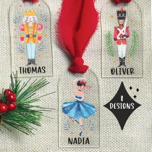 May include: Clear acrylic Christmas ornaments with festive designs. The ornaments feature a nutcracker, a soldier, and a ballerina, each with a name printed below. A red ribbon is attached to the top of the ornaments. The bottom right ornament has the text "8 DESIGNS".