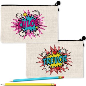 Puede incluir: Dos estuches rectangulares de lona beige con cremallera y gráficos estilo cómic. Uno muestra el nombre CHLOE en letras rosas dentro de una estrella azul, y el otro muestra TREVOR en letras azules dentro de una estrella amarilla.