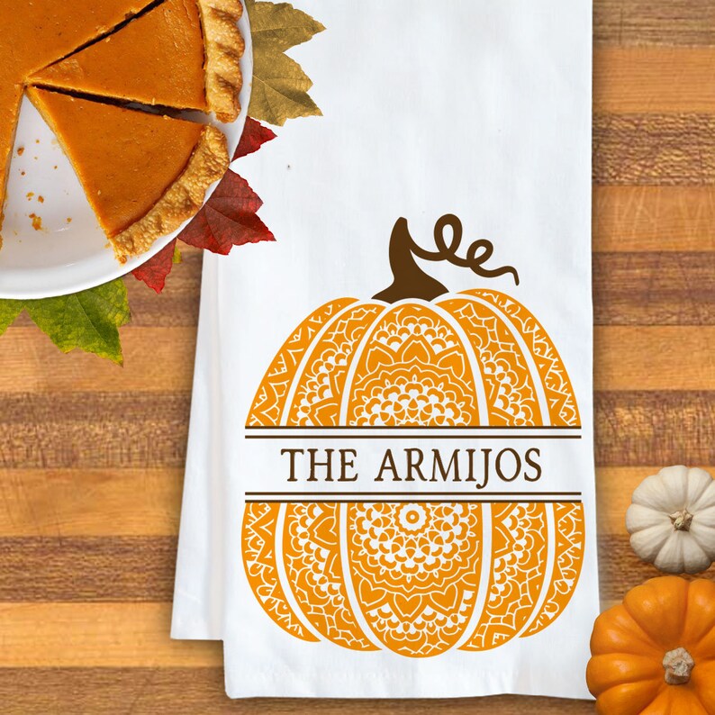 May include: A white kitchen towel featuring a large orange pumpkin graphic with intricate mandala-style patterns. A brown horizontal band across the centre contains the text THE ARMIJOS in a dark brown serif font, topped with a brown pumpkin stem.