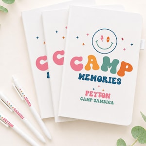 May include: Three white notebooks with a colorful graphic featuring a smiley face, stars, and the text CAMP MEMORIES, PEYTON, and CAMP SAMBICA. Three white pens are included, each personalized with names like JENNA, CASSIDY, and PEYTON in colorful fonts.
