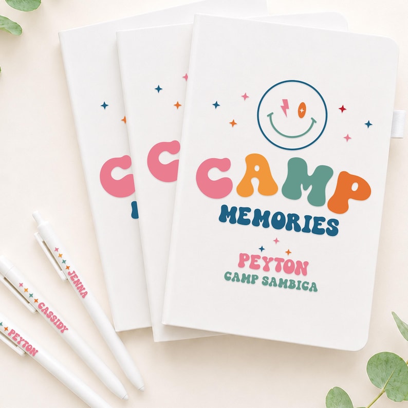 May include: Three white notebooks with a colorful graphic featuring a smiley face, stars, and the text CAMP MEMORIES, PEYTON, and CAMP SAMBICA. Three white pens are included, each personalized with names like JENNA, CASSIDY, and PEYTON in colorful fonts.