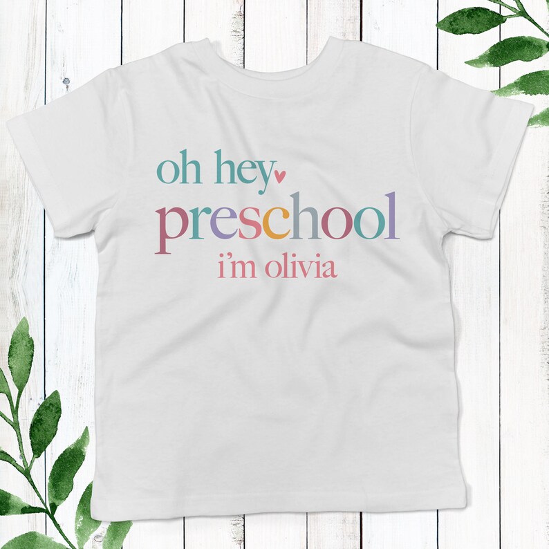 May include: White t-shirt with the text "oh hey preschool i'm olivia" in colorful letters. The text is in a playful font and includes a small pink heart.