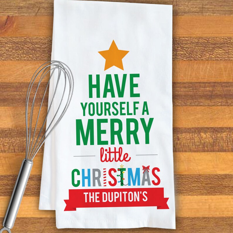 May include: White kitchen towel with a green Christmas tree design. The tree is made up of the words "Have Yourself a Merry little Christmas". The text "The Dupiton's" is in a red banner at the bottom of the tree.