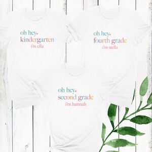 May include: Three white t-shirts with colorful text. The shirts say "oh hey kindergarten i'm ella", "oh hey fourth grade i'm stella", and "oh hey second grade i'm hannah".