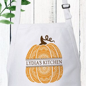 May include: A white apron featuring a large orange pumpkin graphic with intricate mandala-style patterns. A brown horizontal band across the centre of the pumpkin contains the text LYDIA'S KITCHEN in brown capital letters. The apron has adjustable straps.