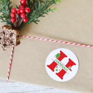 May include: A white round sticker with a red letter H and the name "HANNAH" written in green. The letter H is split in half with a Christmas tree on the top half and Christmas ornaments on the bottom half.