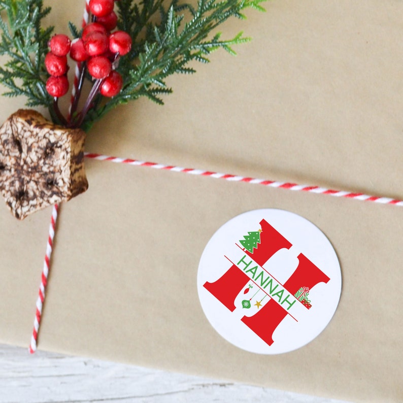 May include: A white round sticker with a red letter H and the name "HANNAH" written in green. The letter H is split in half with a Christmas tree on the top half and Christmas ornaments on the bottom half.
