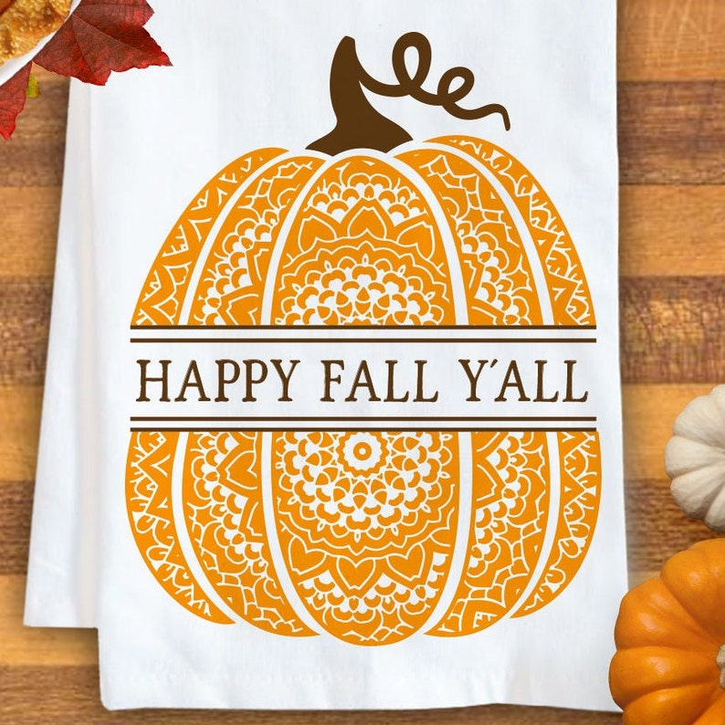 May include: A white kitchen towel featuring a large orange pumpkin graphic with intricate white mandala-style patterns. A brown stem sits atop the pumpkin, and the text HAPPY FALL Y'ALL is printed in brown letters across a horizontal centre bar.