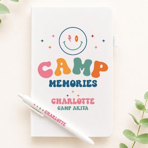 May include: A white notebook and a matching white pen. The notebook cover features a smiley face with a lightning bolt eye, the text "CAMP MEMORIES", "CHARLOTTE", and "CAMP AKITA" in colorful, wavy fonts, surrounded by small star icons.