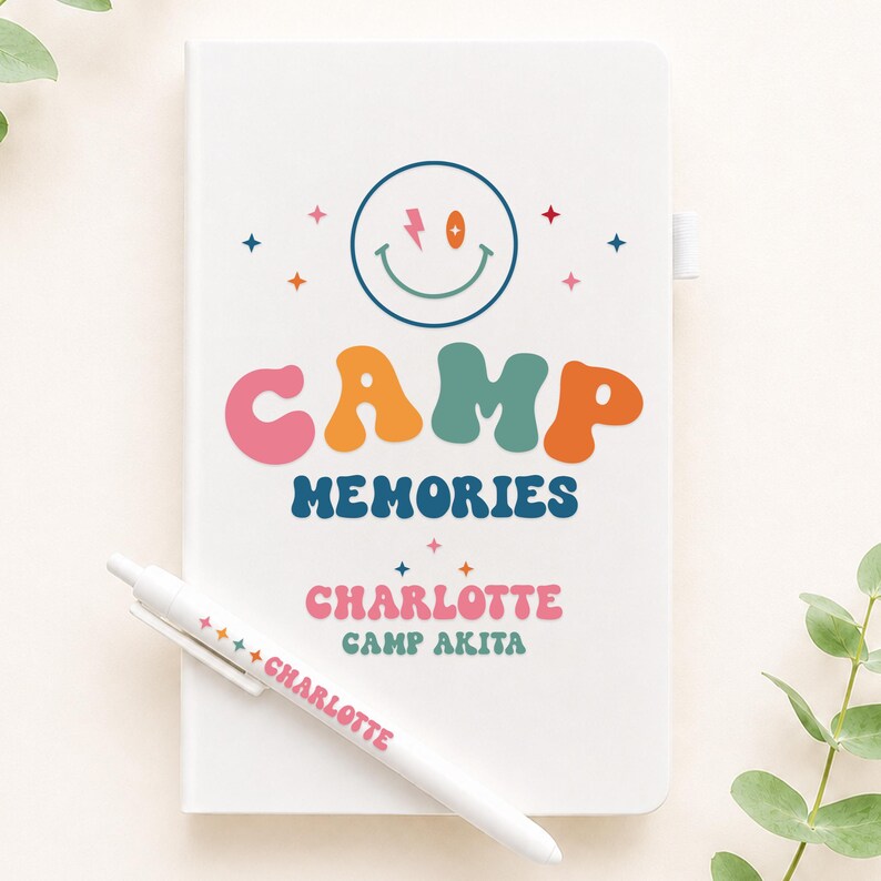 May include: A white notebook and a matching white pen. The notebook cover features a smiley face with a lightning bolt eye, the text "CAMP MEMORIES", "CHARLOTTE", and "CAMP AKITA" in colorful, wavy fonts, surrounded by small star icons.